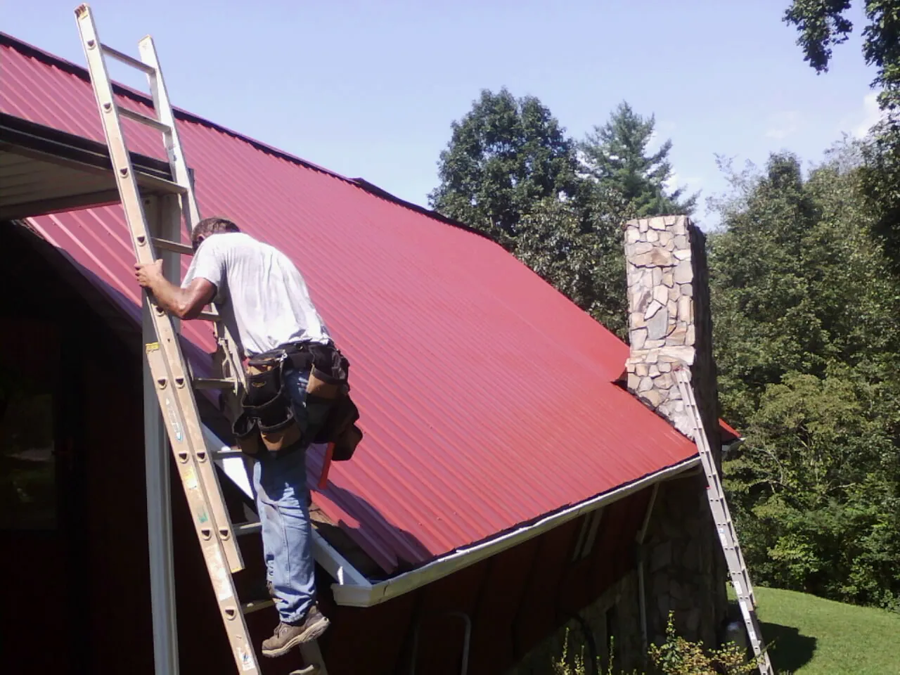 Roofer on a ladder beside a red metal roof and stone chimney — Soffit & Fascia Repair by Roofing Brothers in Everson